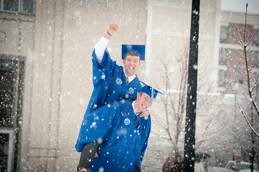 Two white male students in blue commencement cap and gown celebrate their graduation during a heavy winter snowfall outside of Federal Hall